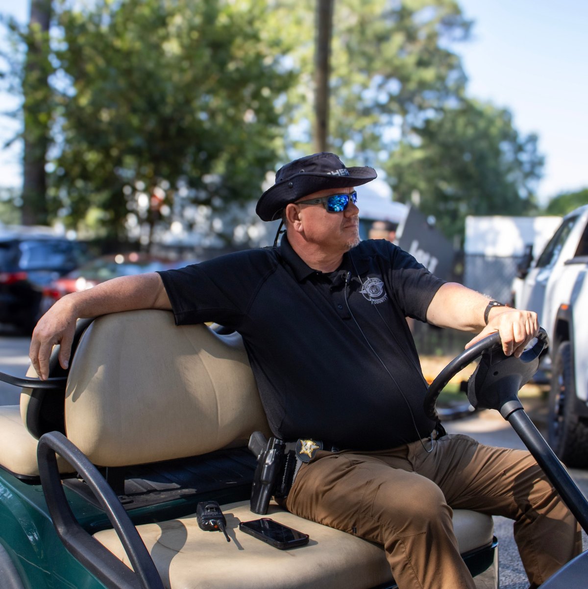 Man in police uniform sitting on golf cart wearing sunglasses and hat in outdoor parking lot