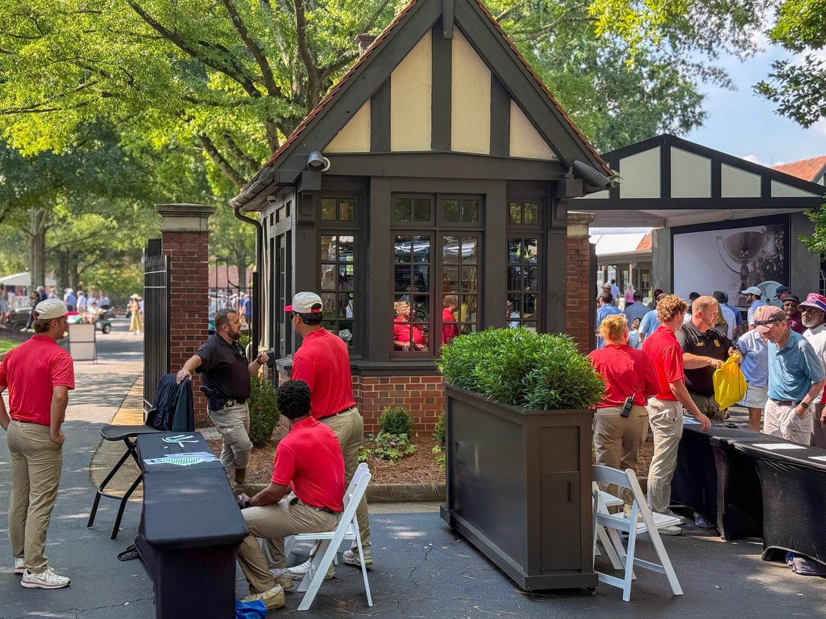Outdoor event with volunteers in red shirts staffing a Tudor-style booth on a tree-lined street with crowds in background