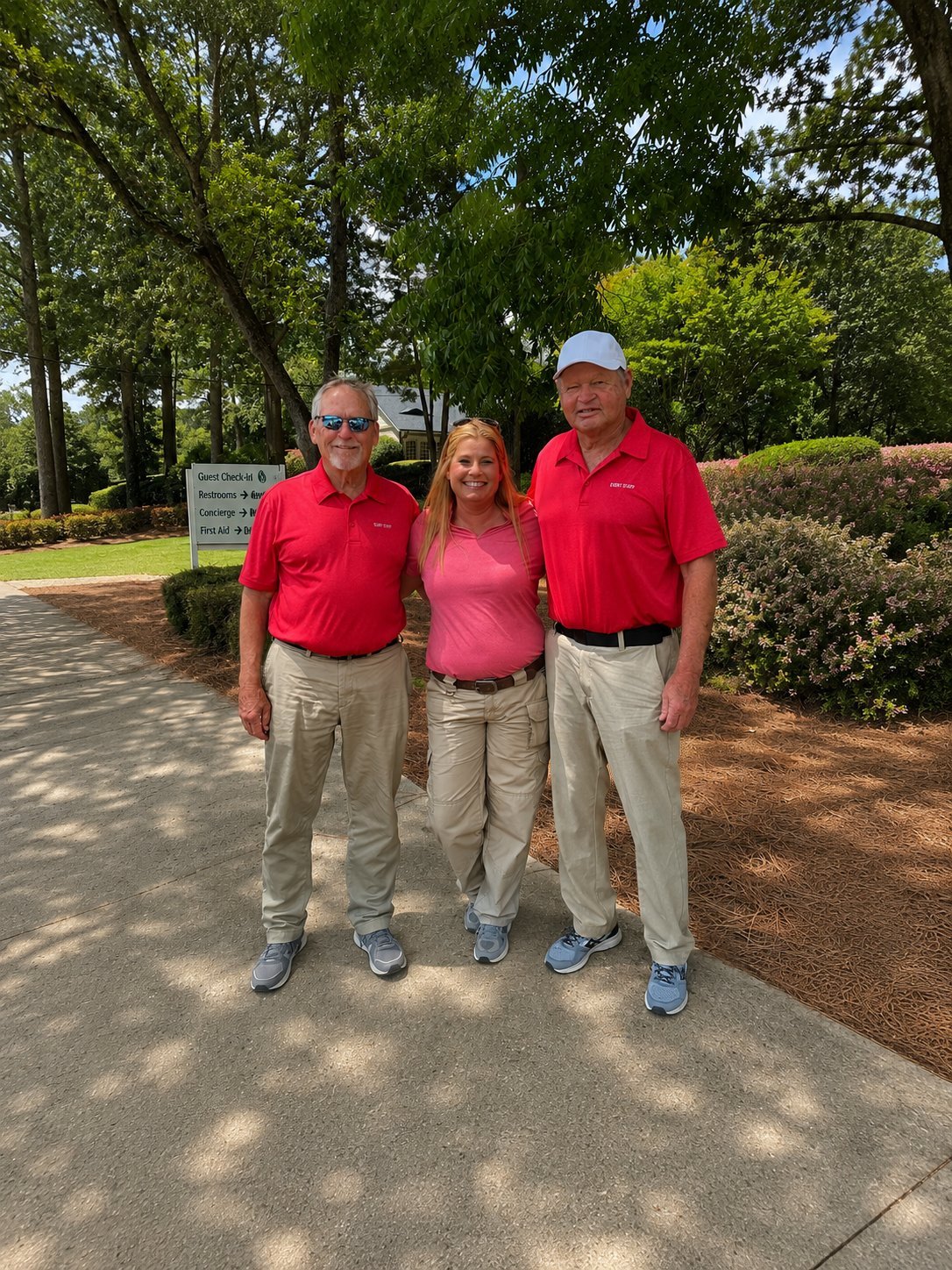 Three people in red shirts and khaki pants standing together on a paved walkway with trees and landscaping in the background