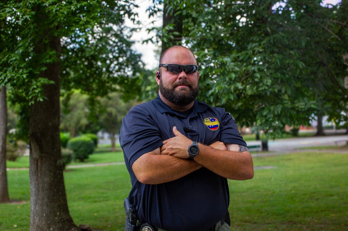 Man with sunglasses and beard wearing dark polo shirt standing in park with arms crossed, trees in background