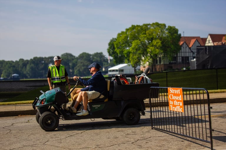 Workers on a motorized cargo scooter next to a construction barrier at a waterfront site with trees and buildings in background