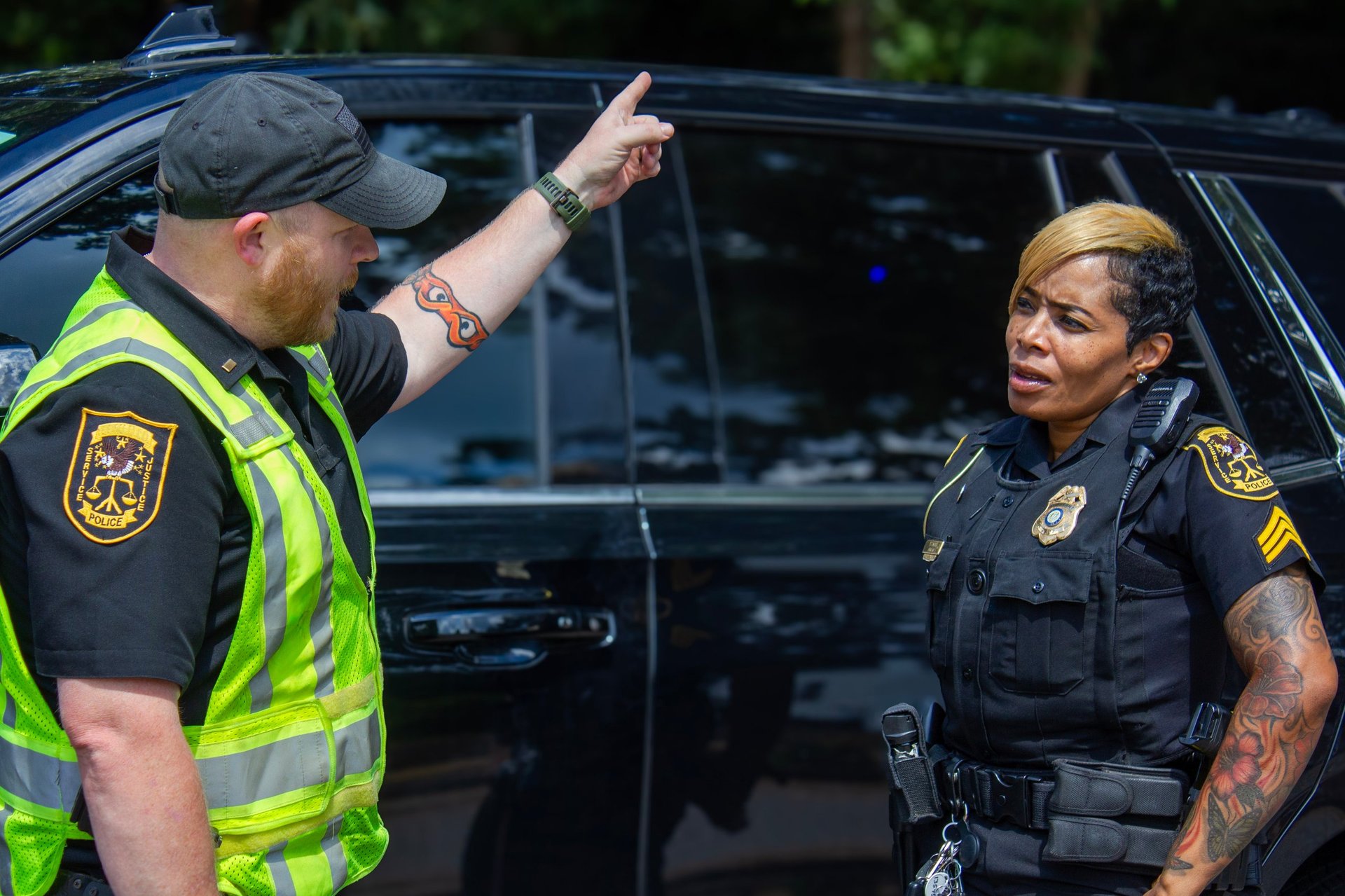 Two police officers in uniform conversing beside a black vehicle, one wearing a high-visibility vest and the other with visible tattoos