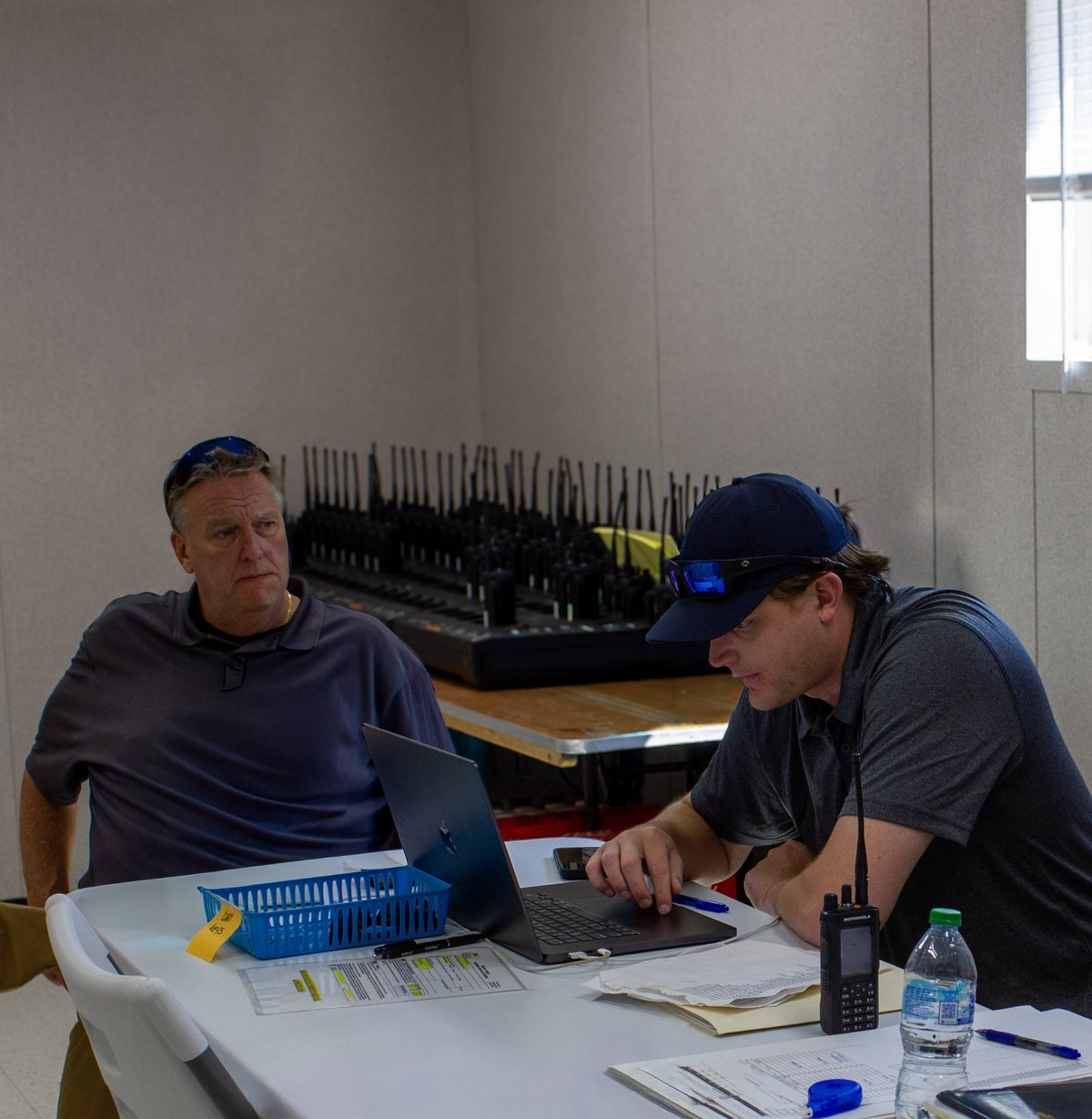 Two workers at a table reviewing documents and using laptops, with industrial equipment visible in the background.