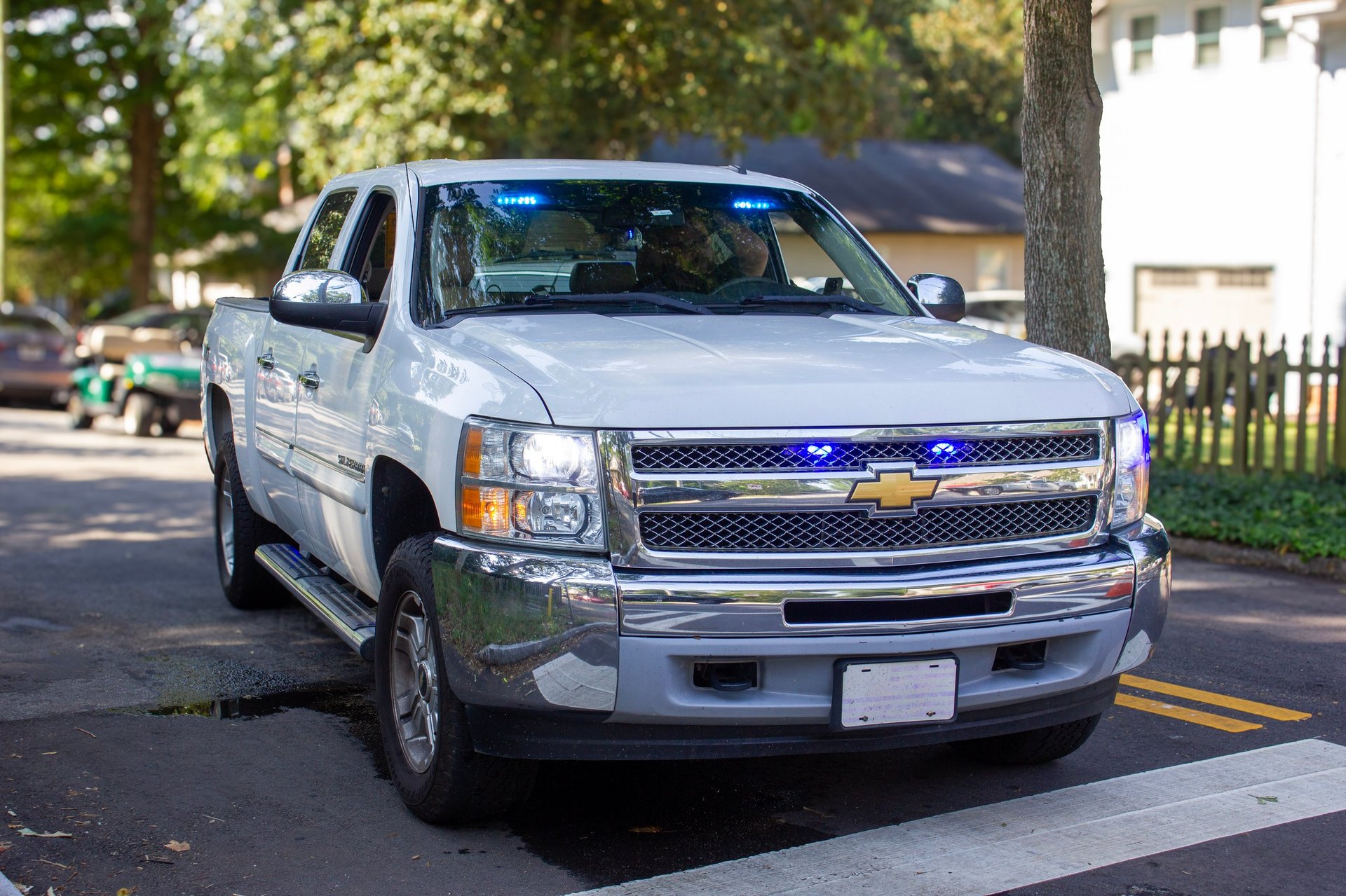White Chevrolet Silverado pickup truck parked on a residential street with trees and white picket fence in background
