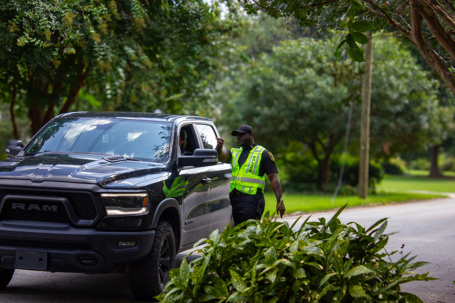 Law enforcement officer in high-visibility vest standing beside black pickup truck on tree-lined road