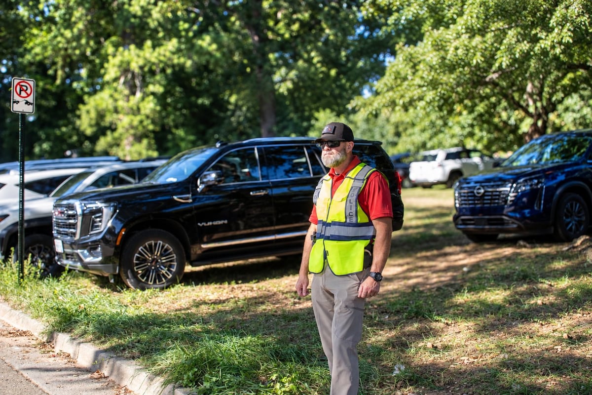 Parking lot attendant in safety vest standing near black SUV with other vehicles in background on sunny day