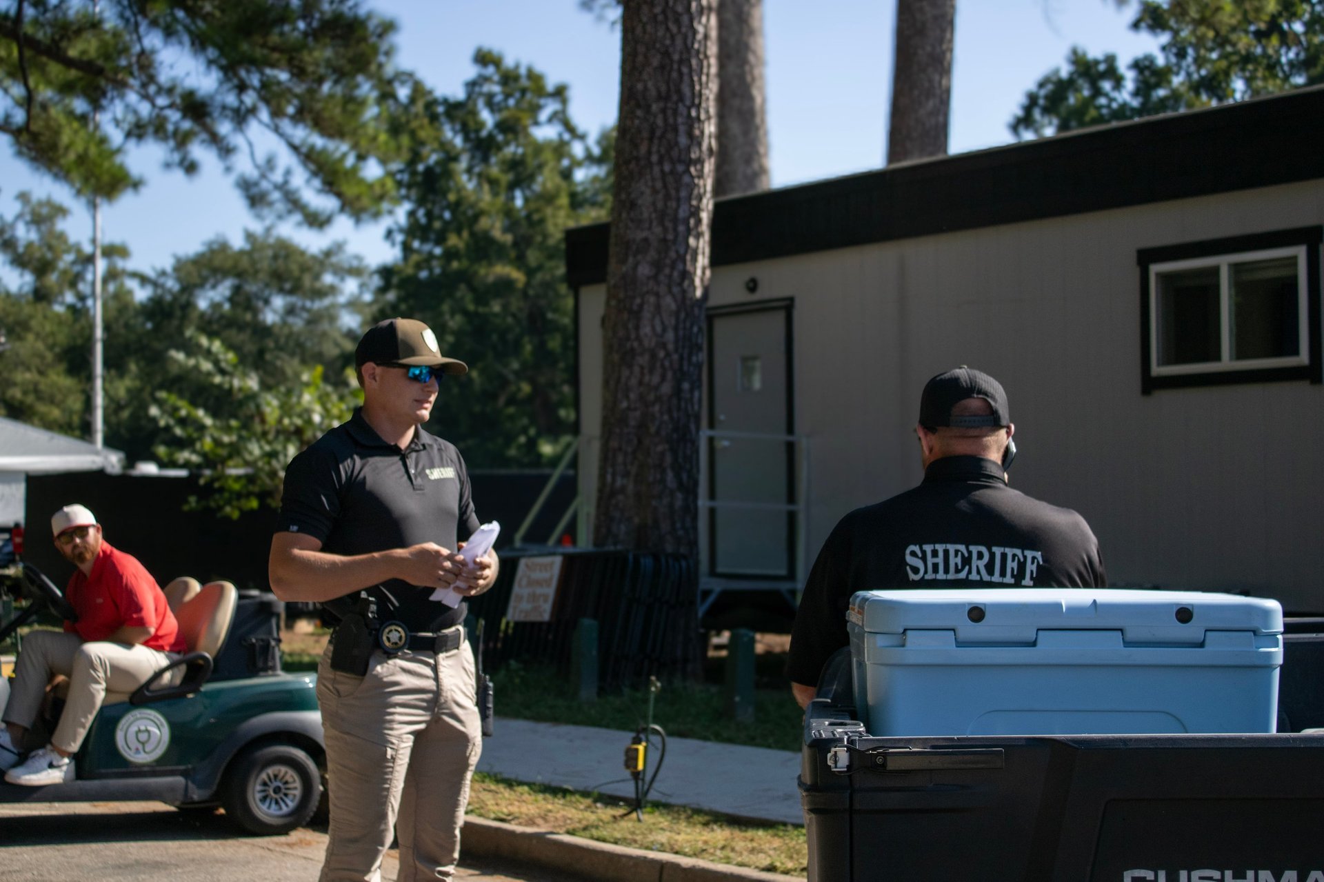 Law enforcement officers at an outdoor scene with a mobile command vehicle, parked near tall trees and a black building.