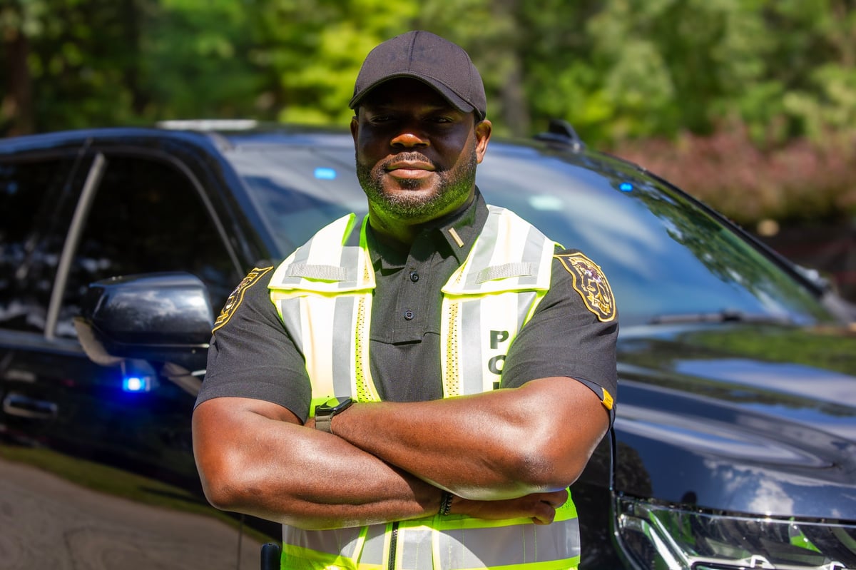 Male security or law enforcement officer in reflective yellow safety vest and cap standing with crossed arms in front of a police vehicle