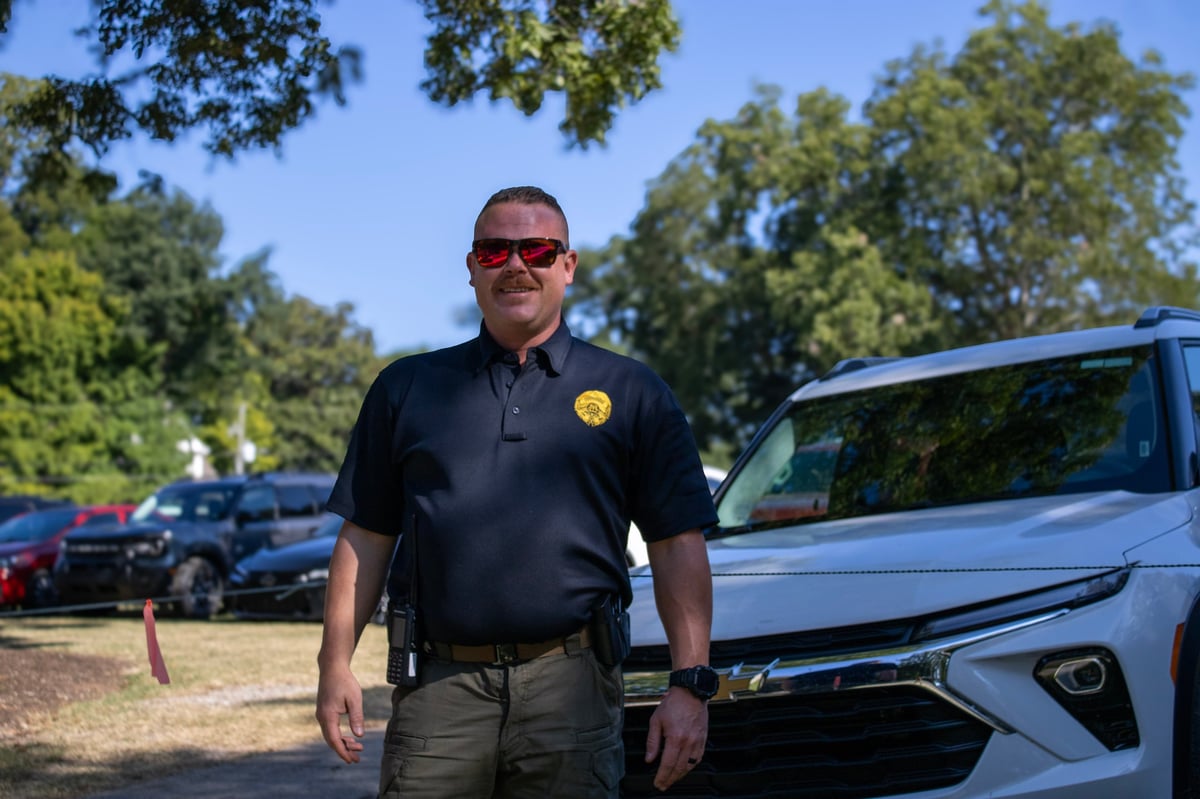 Man in dark polo shirt and sunglasses standing beside a white car in a parking lot with trees in background