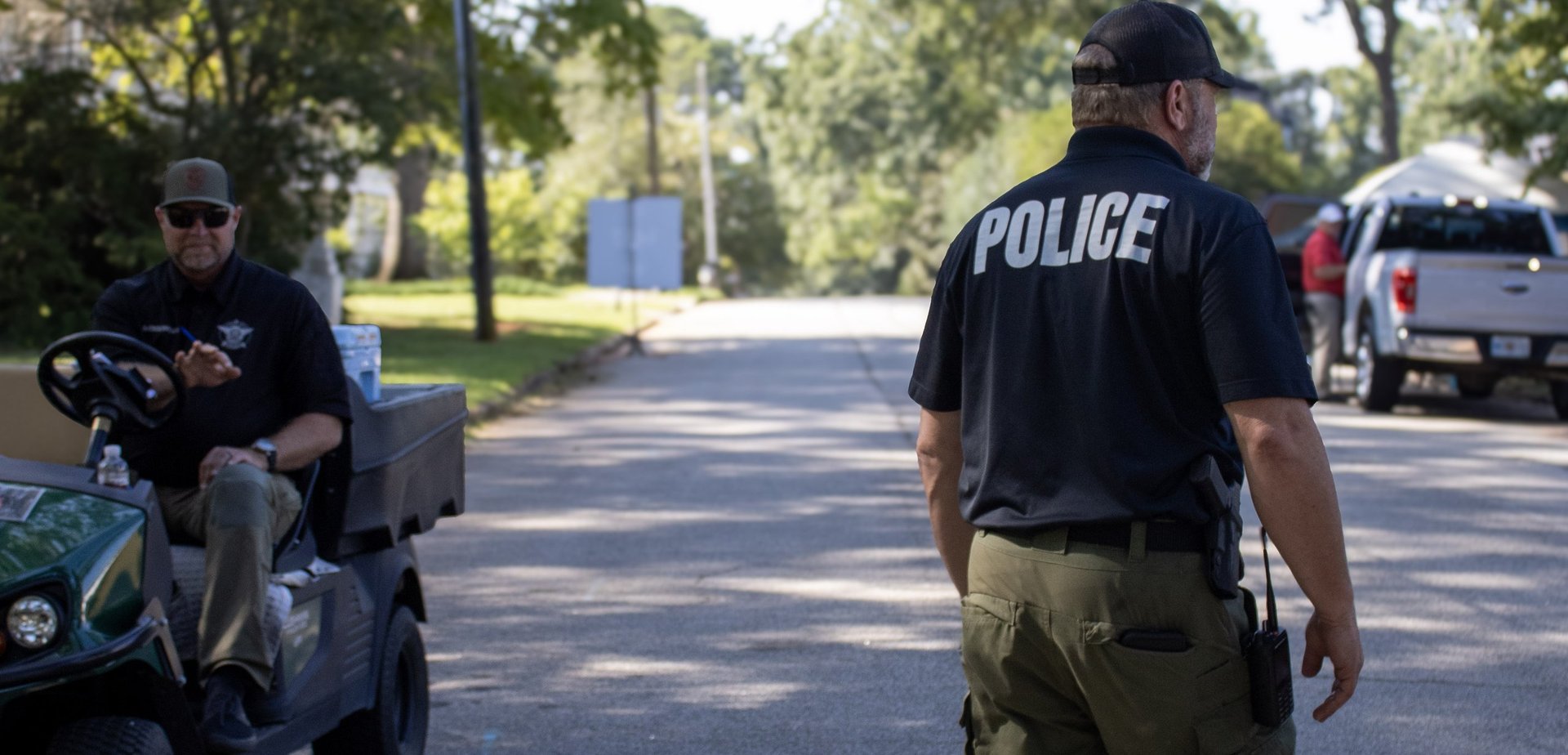 Police officers on a tree-lined residential street with patrol vehicles in the background.