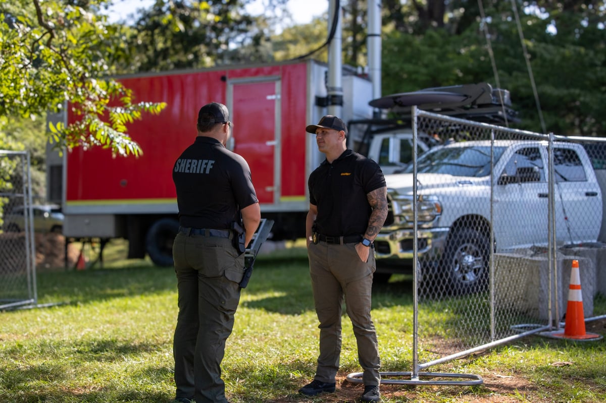 Two sheriff deputies stand beside a chain-link gate with a red mobile command vehicle and white police truck visible in the background.