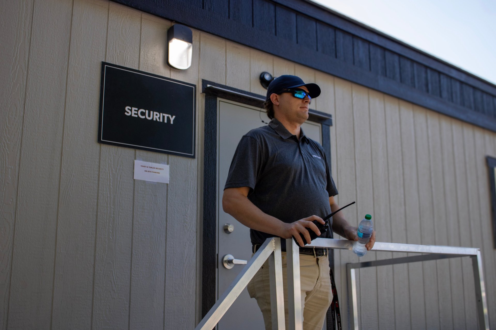 Security guard in black uniform and cap standing on metal platform at industrial building entrance with security sign