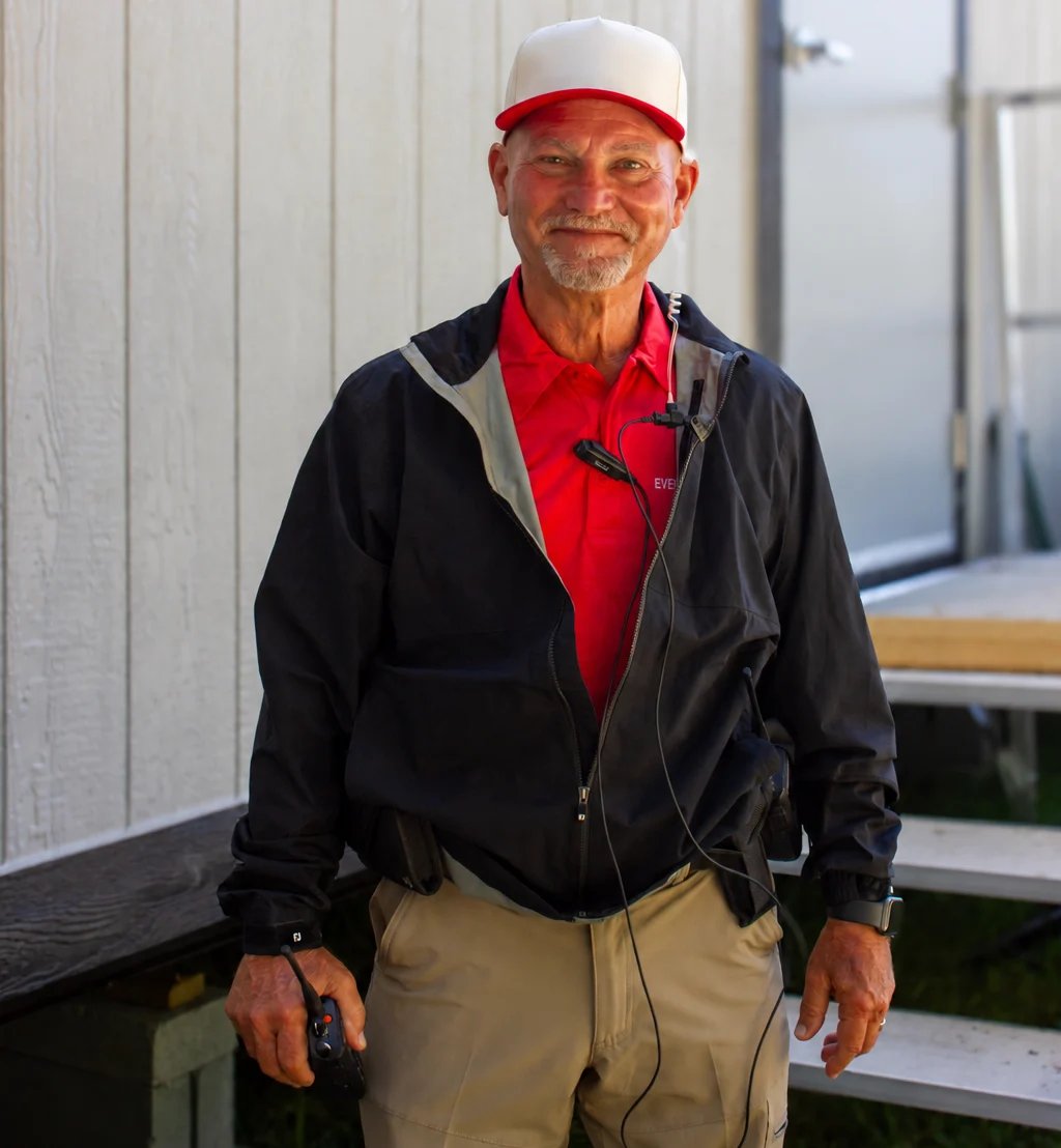 Smiling man wearing red and white cap, black jacket, and beige pants standing in an indoor setting