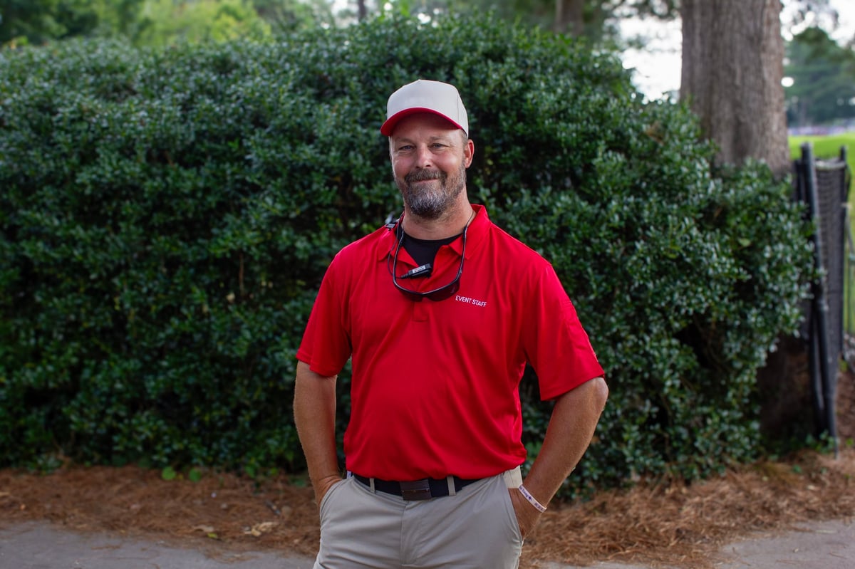 Man in red polo shirt and white cap standing with hands on hips in front of green hedges