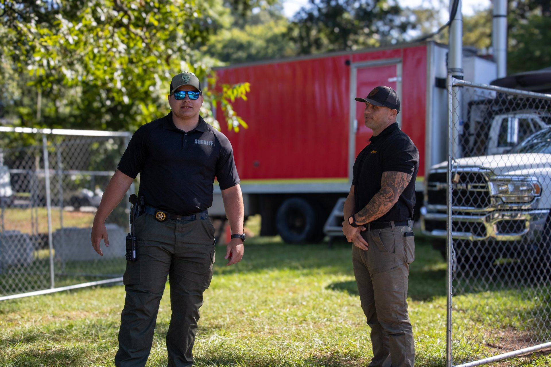 Two uniformed law enforcement officers stand outdoors near a red cargo trailer and chain-link fence