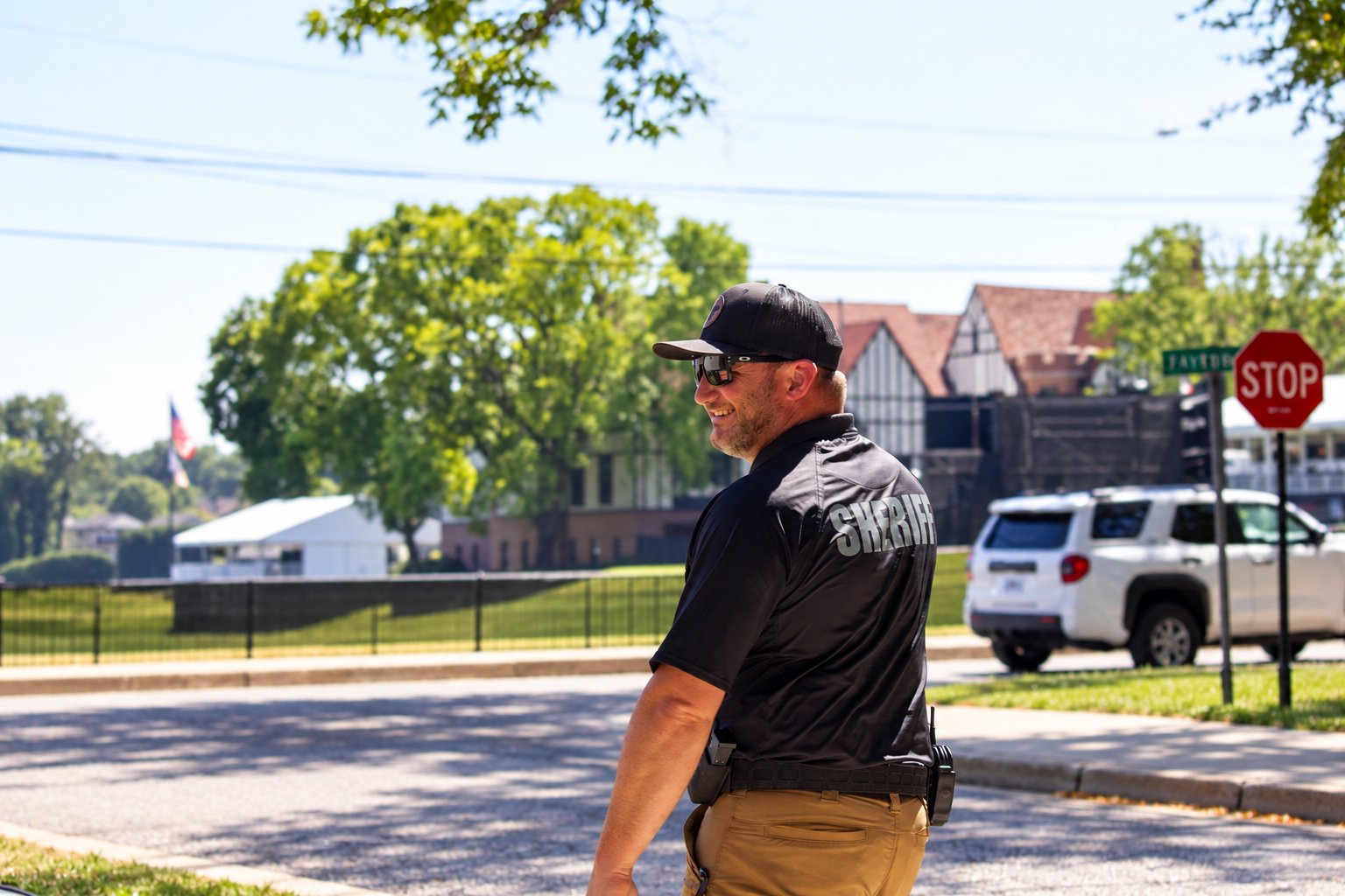 Man in black shirt and baseball cap standing at a residential street intersection near a stop sign, with trees and houses visible in background