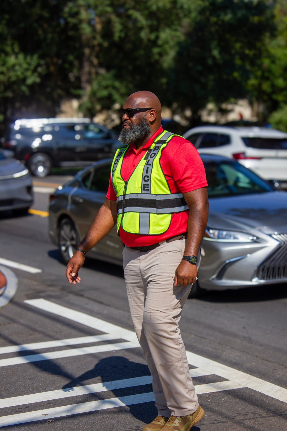 Man in red shirt and neon yellow safety vest walking through parking lot with cars in background