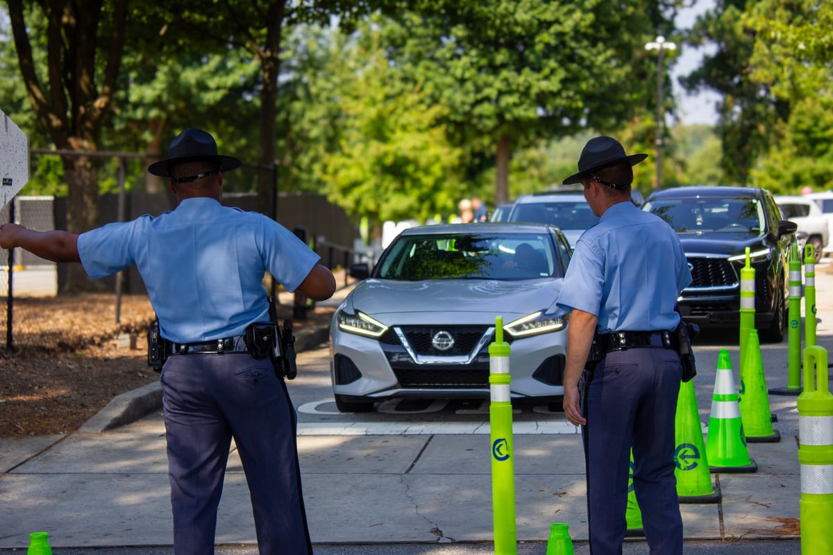Two parking enforcement officers in blue shirts directing traffic in a parking lot with a silver Nissan vehicle and green parking bollards visible