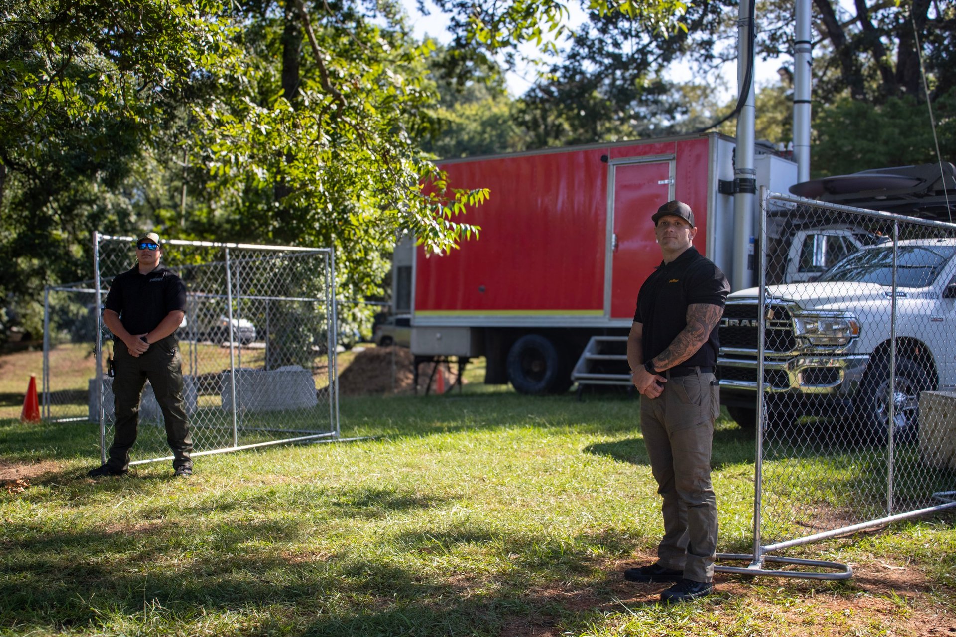 Two men installing or repairing a chain-link fence gate in a grassy yard with a red trailer and trees in the background