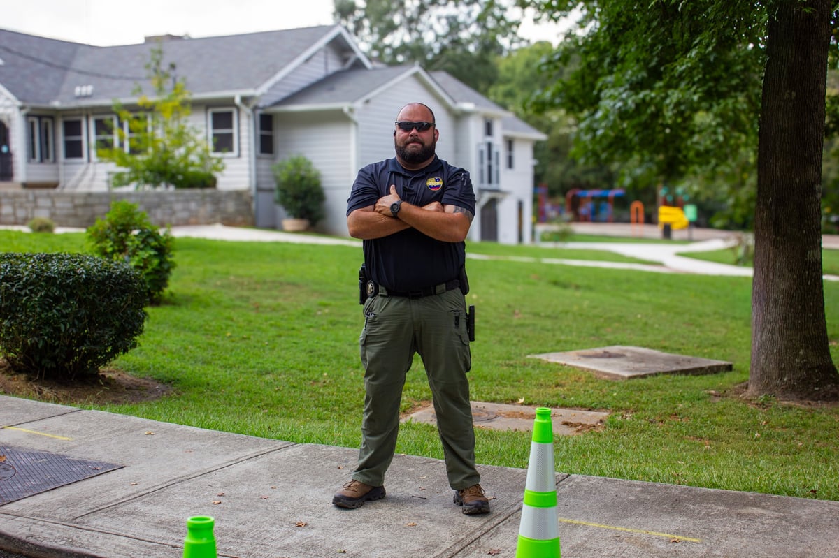 Man in uniform standing with arms crossed on residential driveway with green traffic cones, house and lawn in background