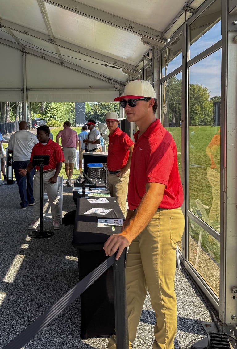 Man in red shirt and white cap standing at a table inside a tent at an outdoor event with green field visible through windows