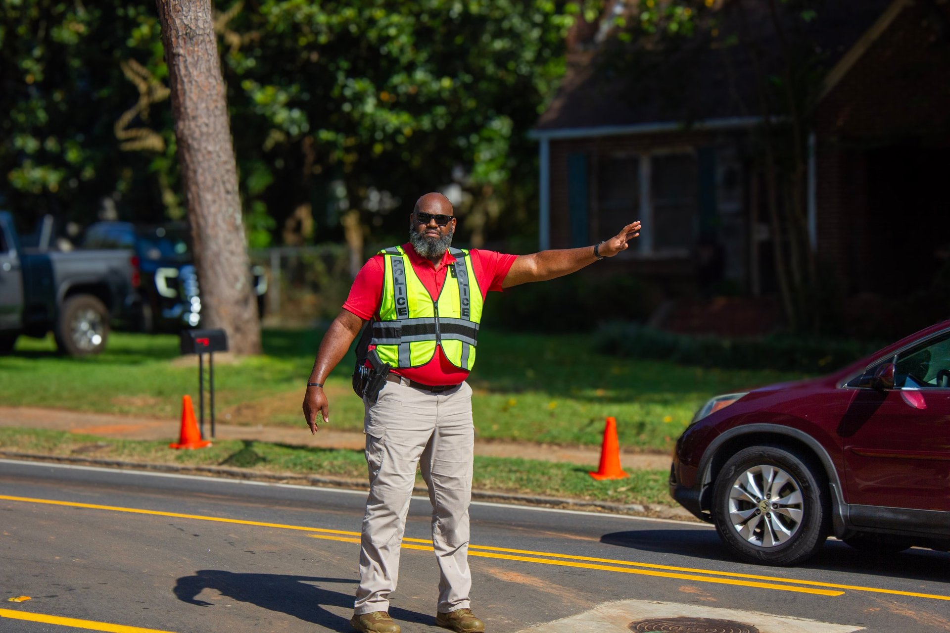 Traffic control worker in red shirt and safety vest directing traffic on residential street with orange cones and parked car