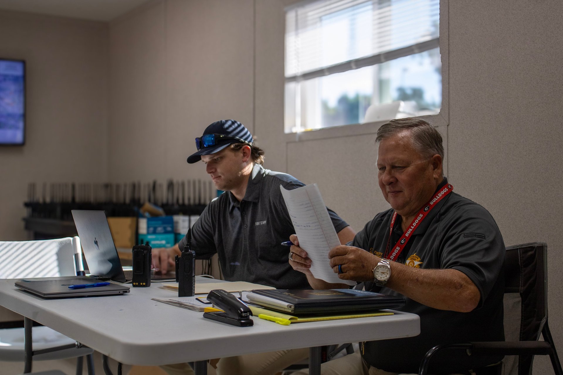 Two men sitting at a table reviewing documents, one wearing a blue helmet and black jacket, the other in a red and black shirt.