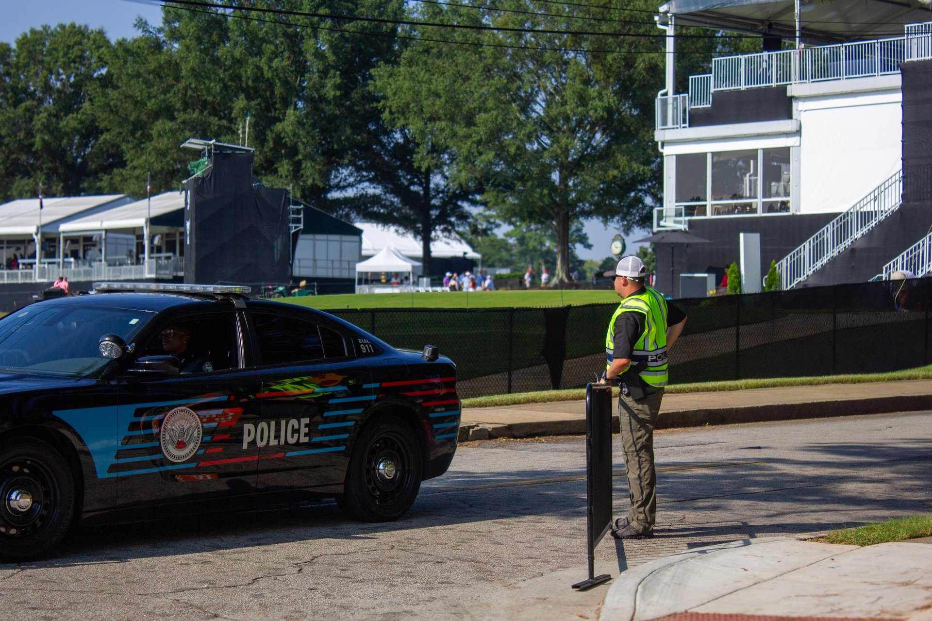 Police officer in yellow vest standing on street next to blue police car with large building visible in background