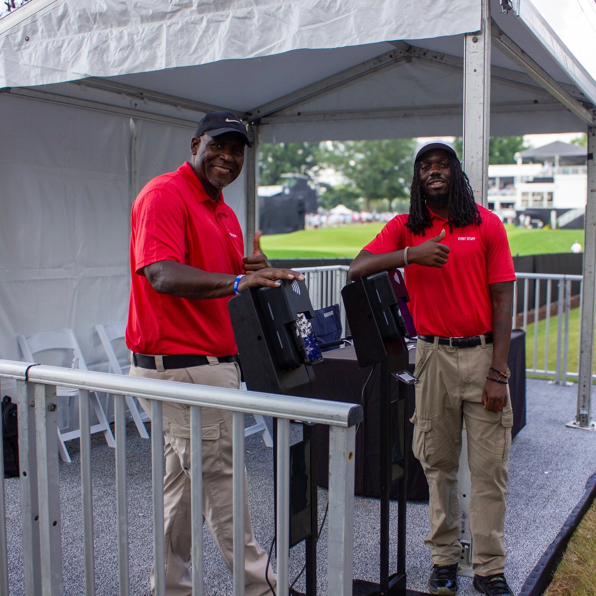 Two men in red shirts holding chairs inside a tent at an outdoor event with a sports field visible in the background