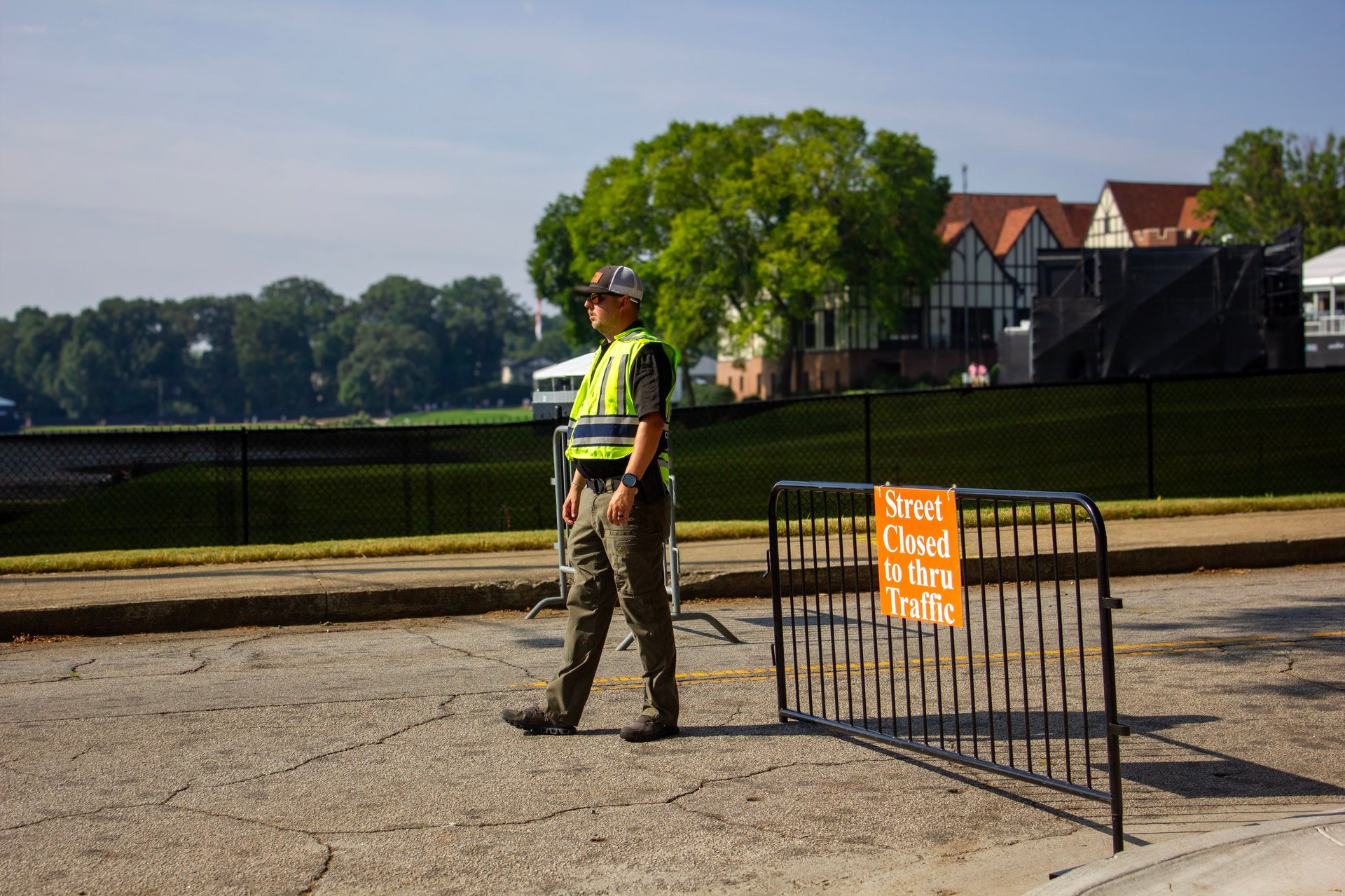 Construction worker in yellow safety vest standing near metal barriers on a waterfront, with trees and buildings visible in background.